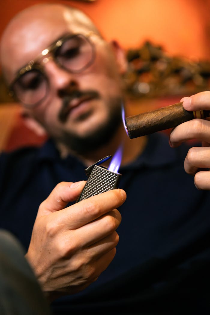 A man wearing eyeglasses lights a cigar with a flame indoors, creating a moody atmosphere.