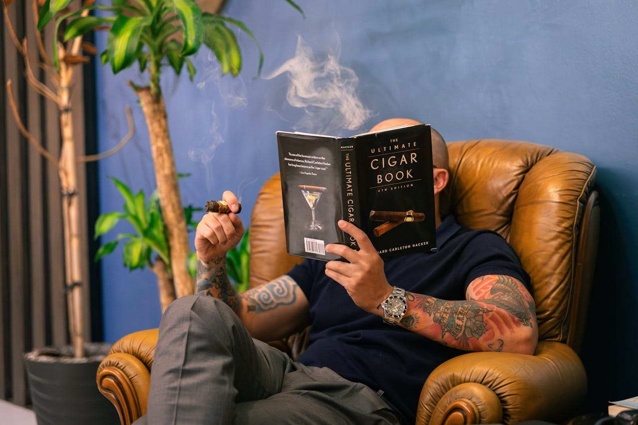 Tattooed man enjoys cigar and book in vintage armchair, creating a relaxed atmosphere indoors.